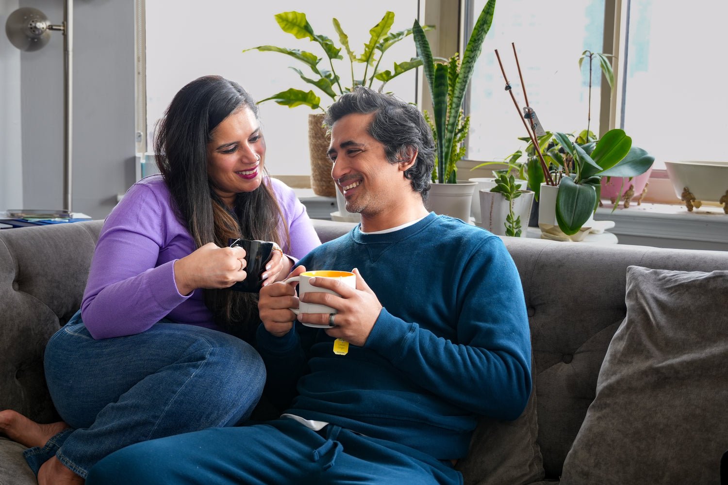 Man and woman sitting on a couch holding mugs, surrounded by plants in a home setting.