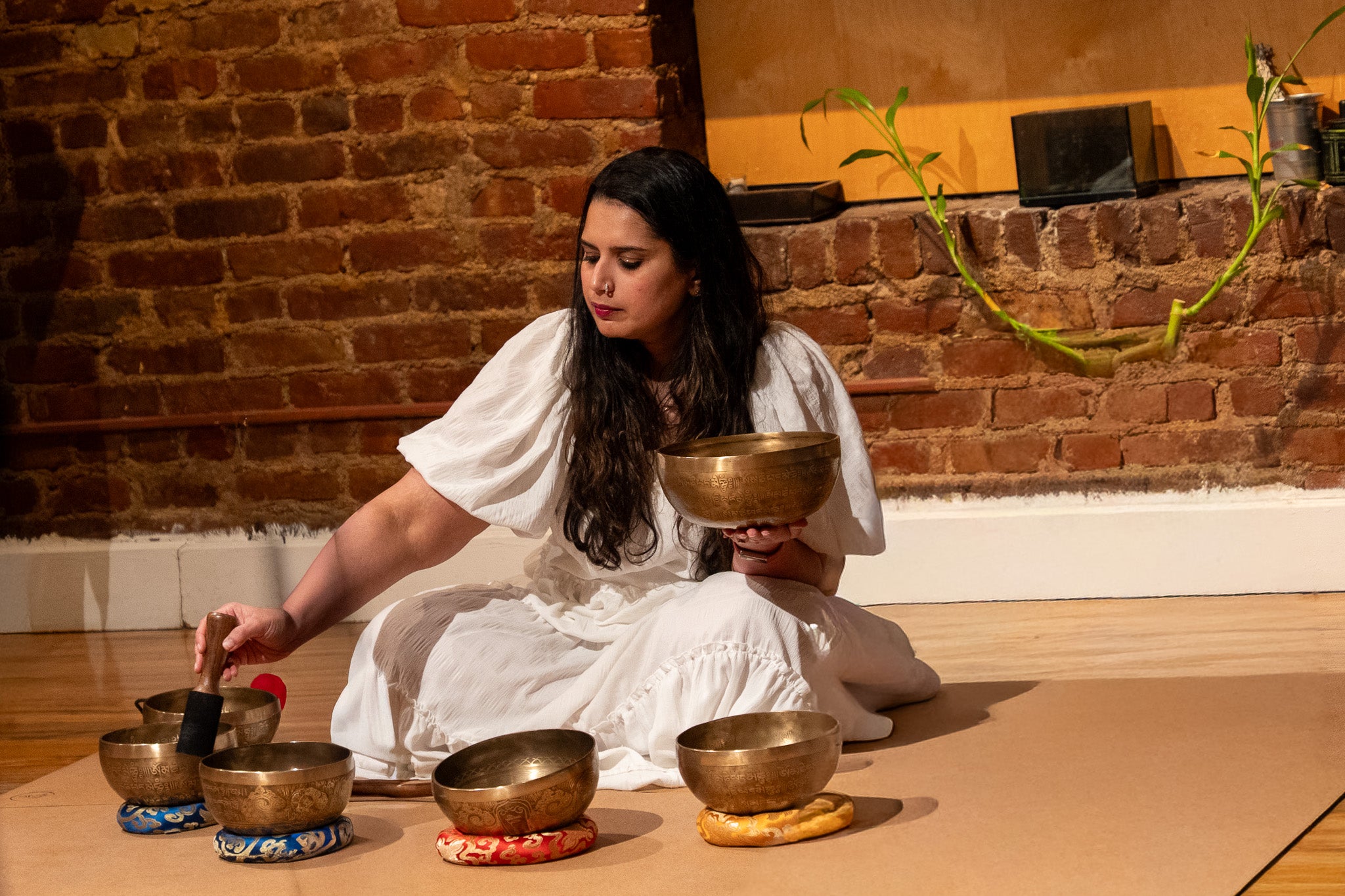 Asmi sitting on the floor with singing bowls in a room with brick walls.